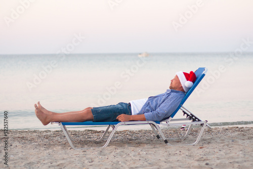 Fototapeta Naklejka Na Ścianę i Meble -  Christmas party, Xmas time, festive . Seasonal holidays. Single young man in Santa hat on chaise-longue on sand of the sea, soft focus, close up view, isolated on blue. Bottle of whiskey in Santa hat