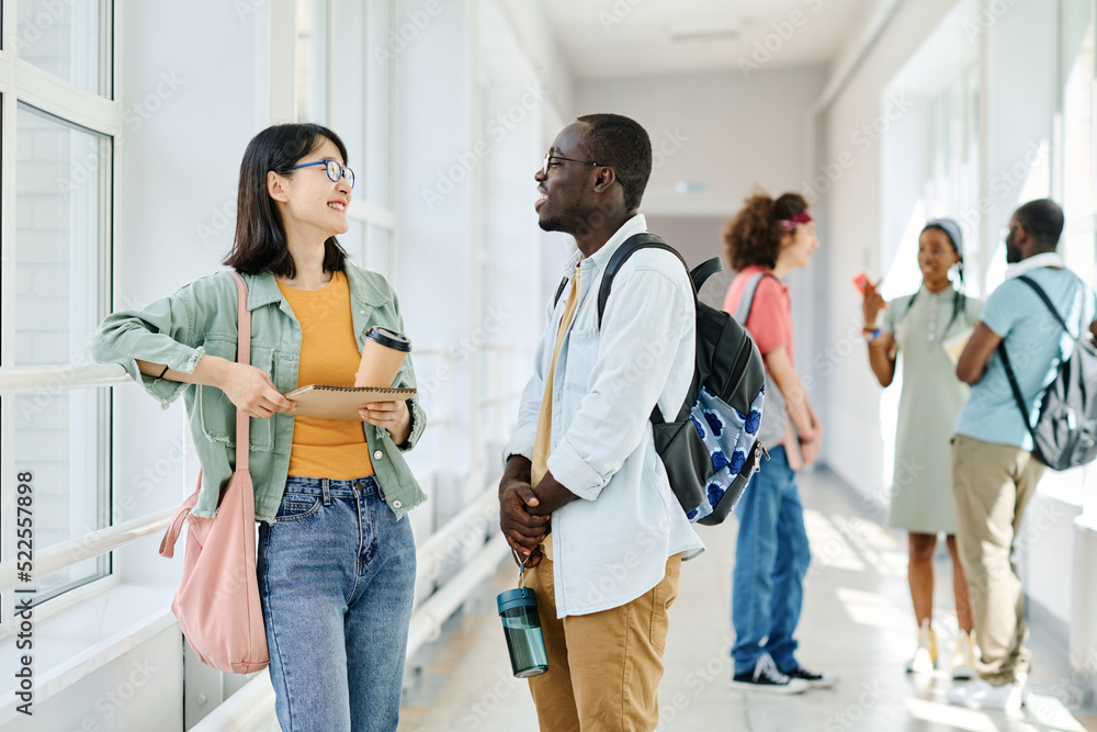 Fototapeta premium Multiethnic students with backpacks chatting during break while standing at school corridor