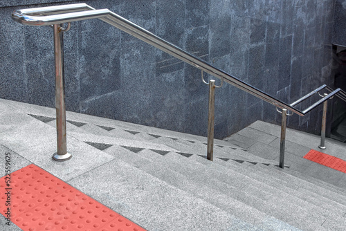 stainless steel iron handrail descent to underground crossing with granite staircase and wall and stone steps, entrance to subway with tactile tiles and ramp for disabled, close-up view, nobody.