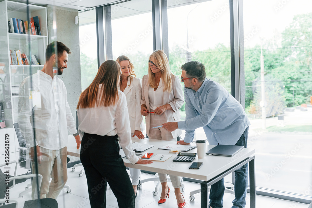 Standing by the table near big windows. Group of professional business people is in the office Standing by the table near big windows. Group of professional business people is in the office