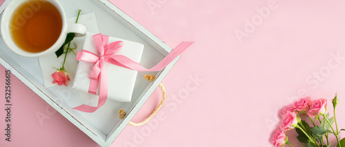 Gift box with tied pink bow and tea in the cup on the white wooden tray on the pink background.Top view. Copy space.