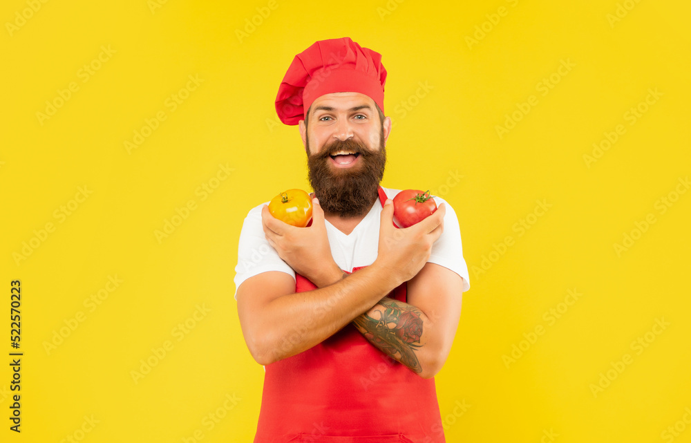 Happy man in cooking apron and toque holding red and yellow tomatoes yellow background, cook