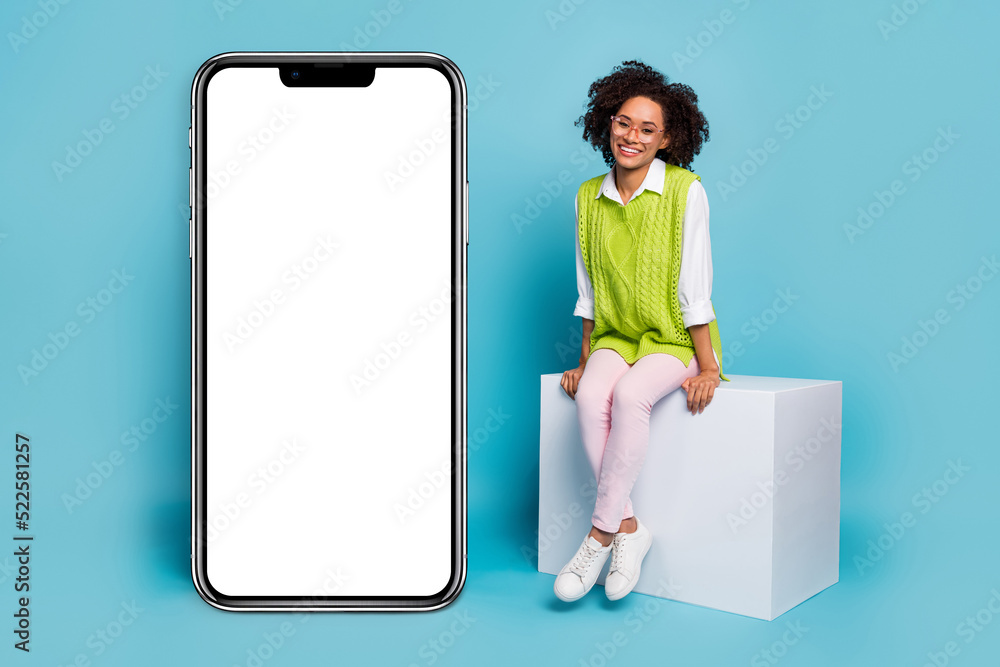 Full size photo of nice millennial brunette lady sit near promo wear eyewear vest pants shoes isolated on blue background Full size photo of nice millennial brunette lady sit near promo wear eyewear vest pants shoes isolated on blue background
