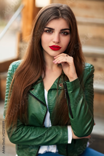Fashion outdoor portrait of gorgeous long hair woman in green leather jacket and red lips, autumn style. Fashionable hipster girl in trendy casual clothes posing at city street lifestyle.