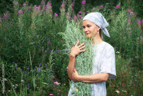 Beautiful village girl hugging bouquet of wormwood (Artemisia absinthium) walks through forest clearing. Slender young woman in headscarf going on flowering lawn in countryside. Lady gathers herbs