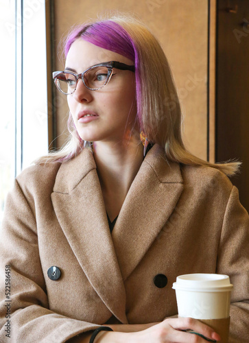 blonde with lilac strands of hair in a beige coat and glasses at a table with a cup of coffee