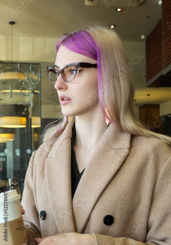 blonde with lilac strands of hair in a beige coat and glasses at a table with a cup of coffee