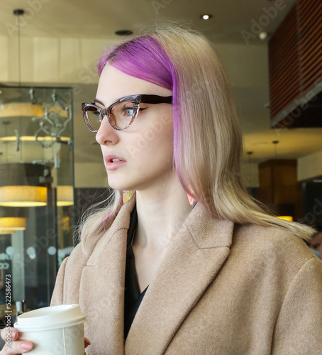 blonde with lilac strands of hair in a beige coat and glasses in a cafe with a cup of coffee