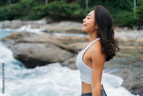 Peaceful and relaxing Asian 30s woman taking a deep breathe in summer beach by seaside. Carefree and happy girl opened arms with relaxation.