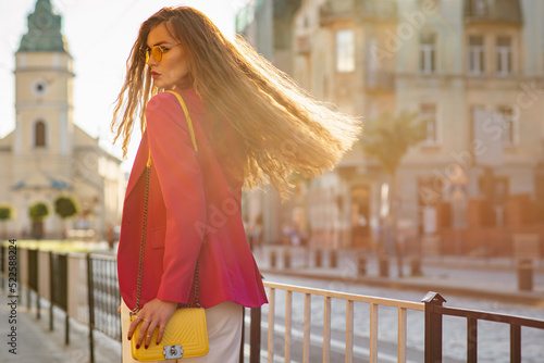 Slika na platnu Fashionable woman with long hair wearing trendy yellow sunglasses, pink fuchsia blazer with faux leather shoulder bag, walking in street of European city