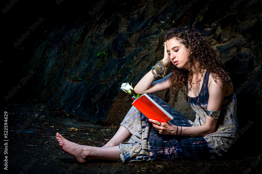 Girl Reading Outside. Wearing long dress, barefoot, a pretty teenage college student with curly long hair is sitting on ground against rocks, holding a red book with a white rose, reading, relaxing..