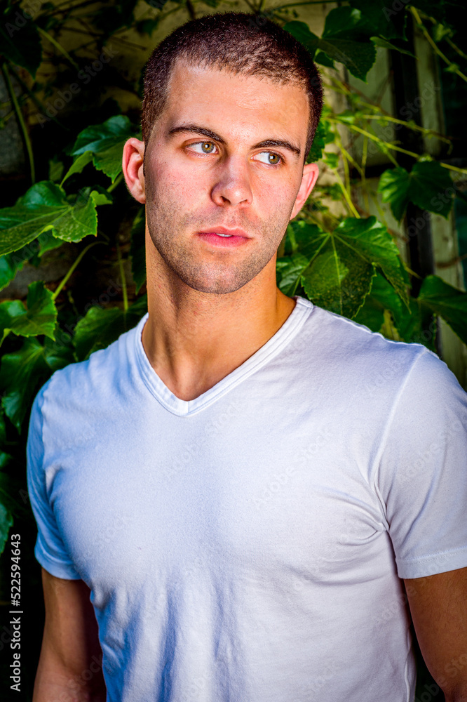 Young man with short hair, wearing white V neck T shirt, standing outdoors by green ivy leavers in New York City, looking away.