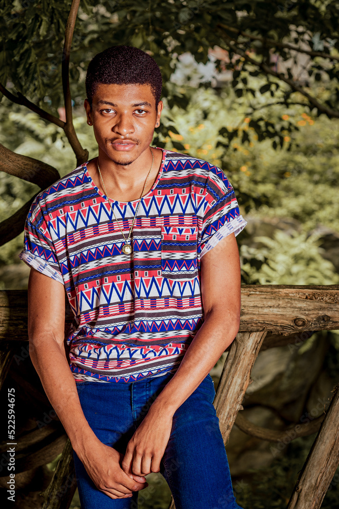 Young African American man wearing colorful pattern shirt, blue jeans, necklace, standing against wooden fence in New York City, charmingly looking forward..