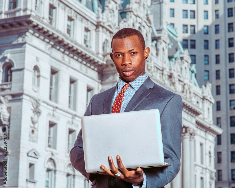 Young African American man wearing blue suit, patterned undershirt, tie, short haircut, standing in business district in New York City, working on laptop computer, thinking.