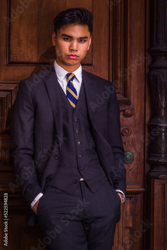 Dressing formally in three pieces black suit, patterned necktie, vest, hands in pockets, a young handsome businessman is standing by an old fashion style office door, looking at you