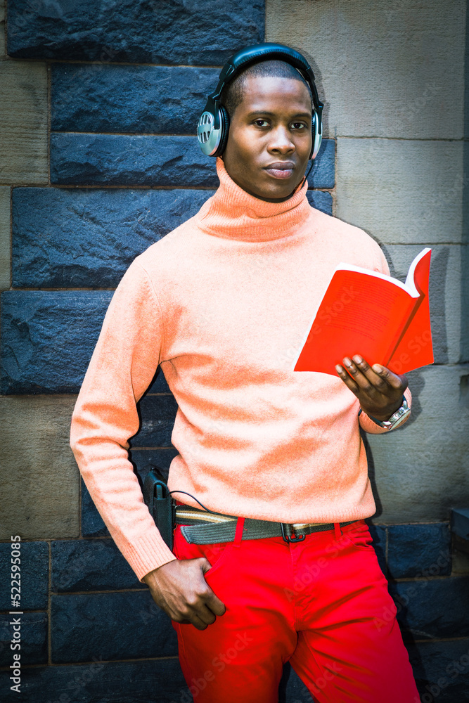 Young man wearing light orange sweater with high collar, red pants, headphones, standing against wall in New York City, reading read book, listening music..