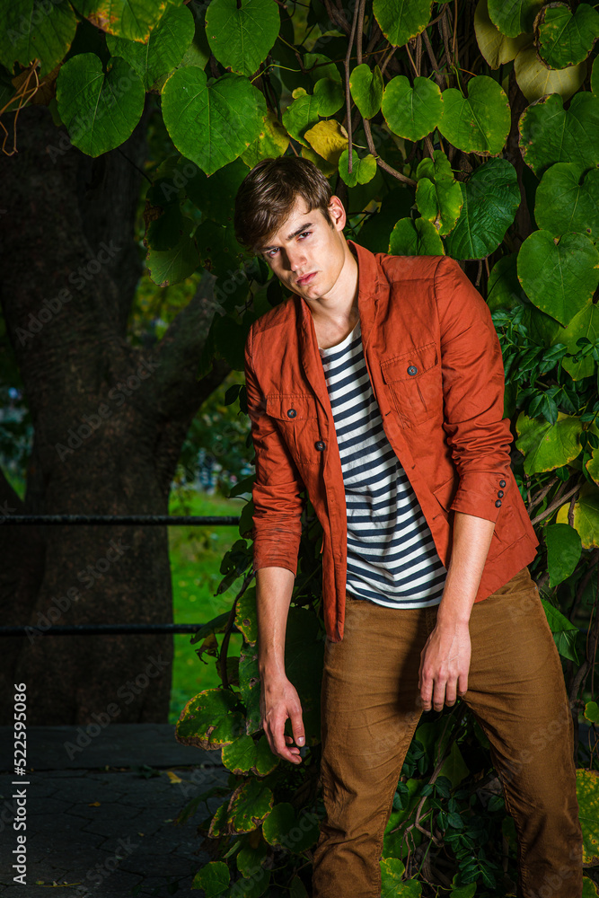 Young Man wearing dark reddish brown jacket, unbuttoned, striped under shit, brown corduroy pants standing by big leaves in New York City, looking at you..