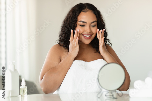 Facelift and skin care concept. Young black plus size woman massaging her face and looking at mirror