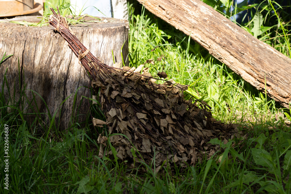 Fototapeta premium Banya broom lying on green grass and rests on stump, warm colors, horizontal photo. Outdoor shot, bath accessory, nature floral and herbal product, health care