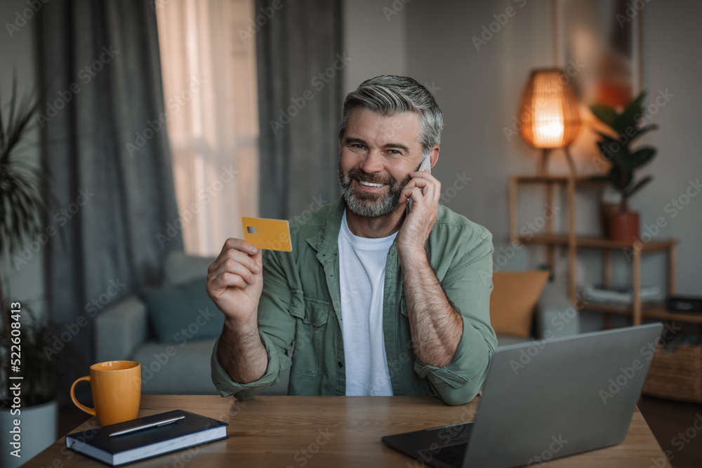 Glad retired caucasian male with beard calls by smartphone, uses credit card and pc to shopping online Glad retired caucasian male with beard calls by smartphone, uses credit card and pc to shopping online