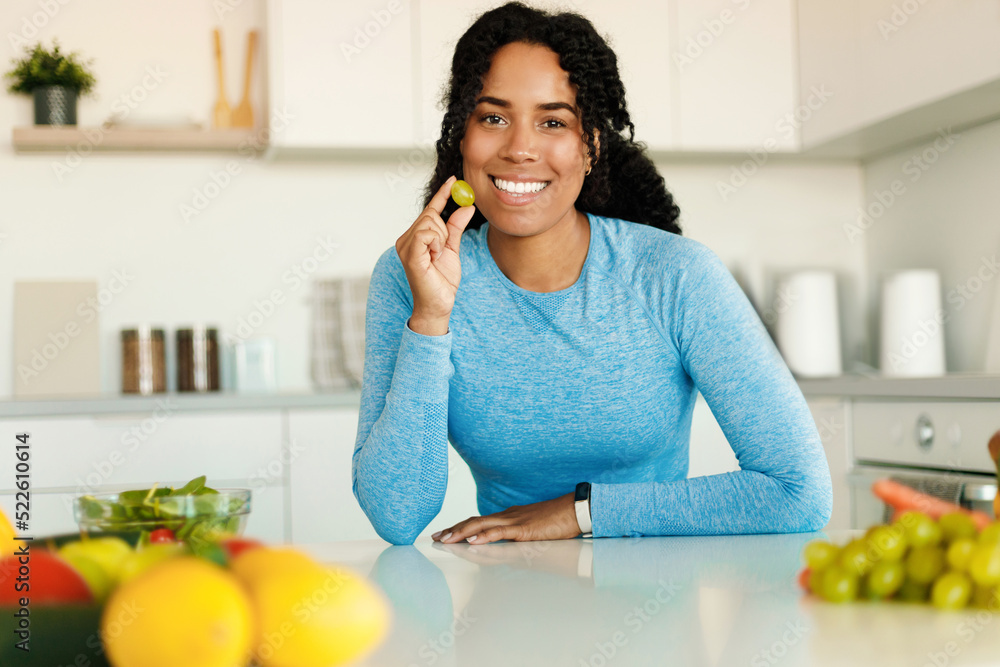 Excited fit african american lady eating fruits for breakfast, holding grape and smiling at camera, sitting in kitchen Excited fit african american lady eating fruits for breakfast, holding grape and smiling at camera, sitting in kitchen