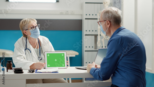 Physician holding horizontal greenscreen on digital tablet  at appointment with senior patient. Chroma key template with isolated copyspace and blank mockup background. Tripod shot.