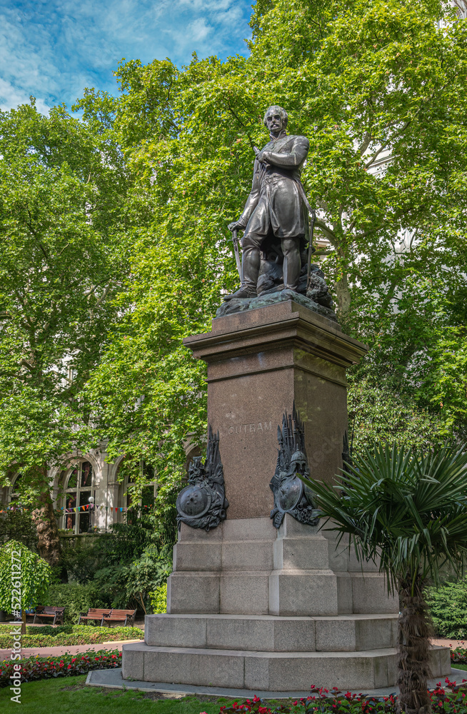 London, UK- July 4, 2022: Closeup of Sir James Outram statue in Whitehall Gardens surrounded by green foliage under small corner of blue cloudscape.  Red flowers add color.