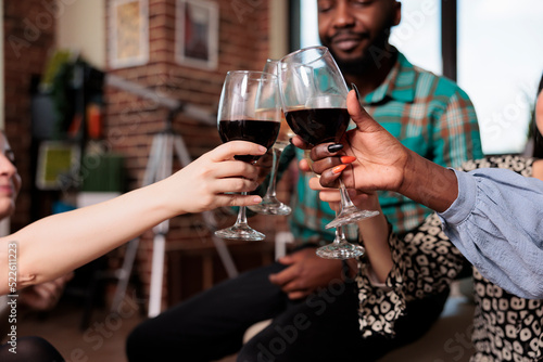 Diverse group of close friends at wine party clinking glasses while enjoying fun time together in living room. Cheerful people toasting wineglasses while celebrating birthday at home.