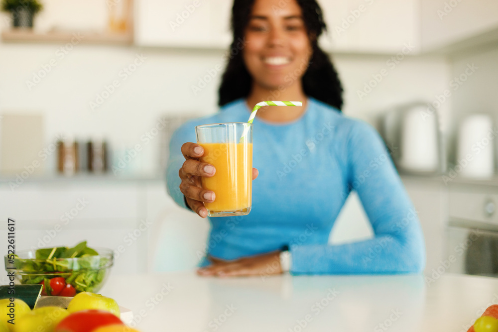 Happy black fitness lady in sportswear showing fresh orange juice in glass, sitting in kitchen after domestic workout, Happy black fitness lady in sportswear showing fresh orange juice in glass, sitting in kitchen after domestic workout,