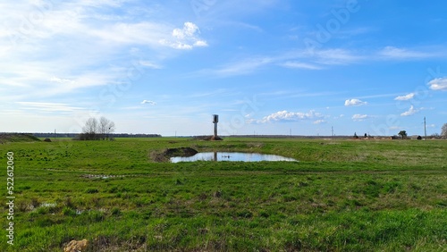 In a grassy meadow not far from the village there is a technical water reservoir and a water tower. Behind the meadow is a forest. It is sunny and the sky is blue with clouds