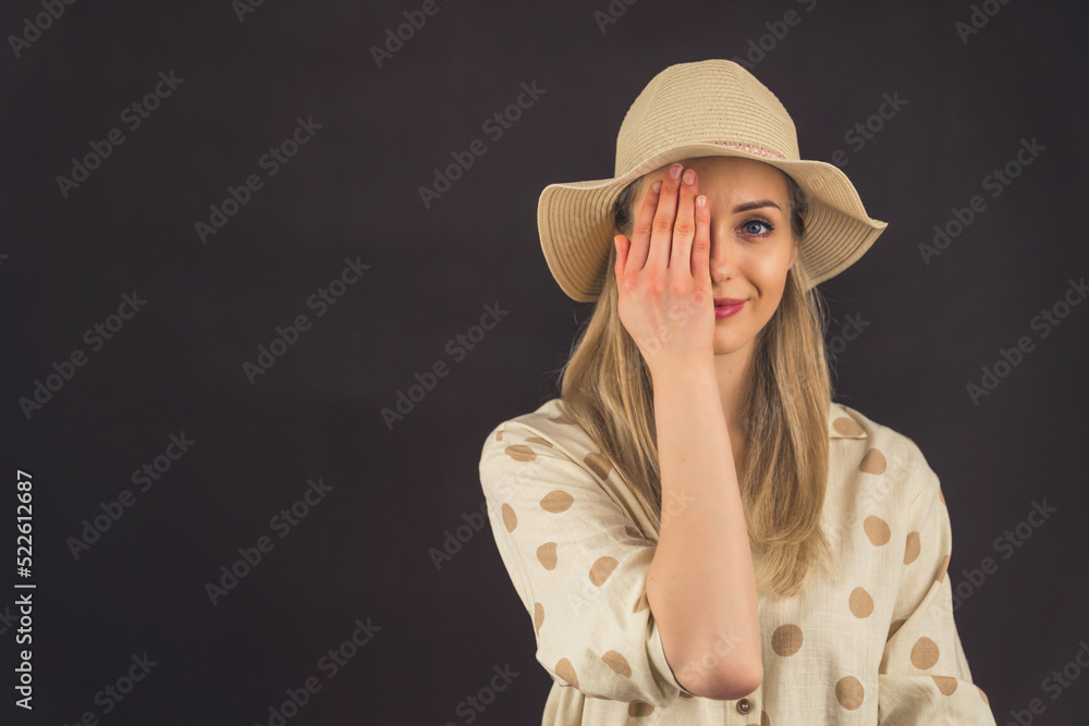 Young white blonde woman wearing linen polka-dot shirt and straw hat covering one eye with hand looking into camera. Dark background studio shot. High quality photo Young white blonde woman wearing linen polka-dot shirt and straw hat covering one eye with hand looking into camera. Dark background studio shot. High quality photo