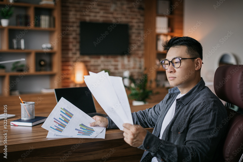 Serious concentrated adult japanese man investor in glasses works with papers, charts and graphs in dark office Serious concentrated adult japanese man investor in glasses works with papers, charts and graphs in dark office