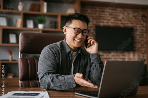 Cheerful mature japanese man in glasses calls by phone, works with laptop at table in office interior