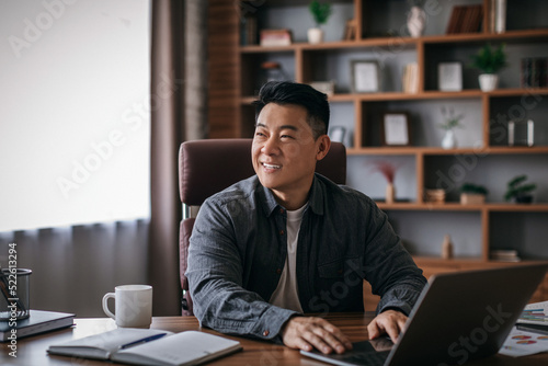 Happy mature chinese man at table working with laptop in home office interior and looking at empty space