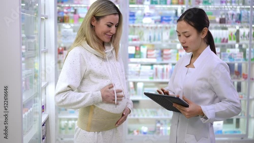 Pregnant woman putting on maternity belt talking with pharmacist in drugstore. Beautiful Caucasian and Asian women choosing belly belt in pharmacy standing with digital tablet photo