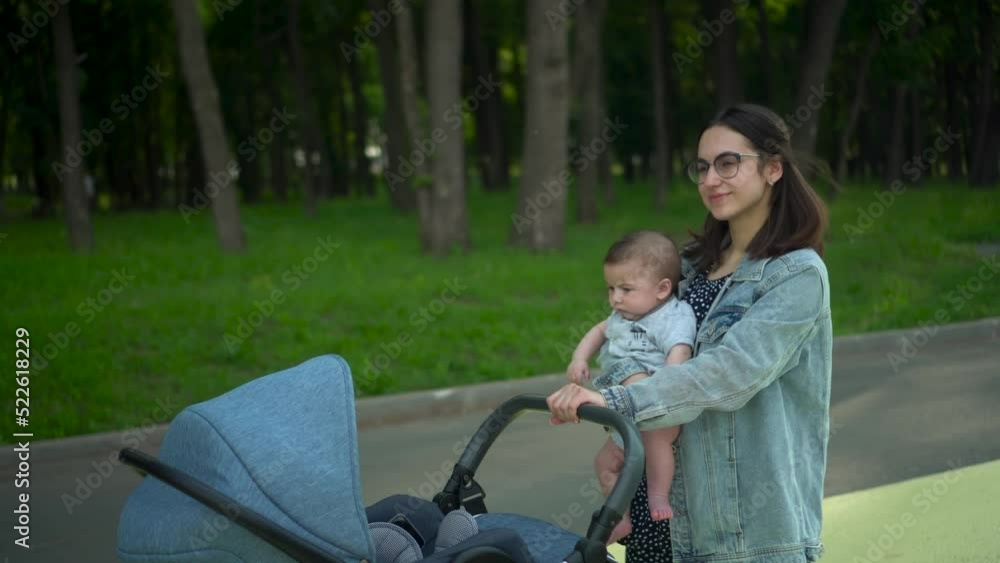 A young mother walks with a six-month-old son carrying him in her arms. A woman with glasses carries a baby and pushes a stroller in the park. A young mother walks with a six-month-old son carrying him in her arms. A woman with glasses carries a baby and pushes a stroller in the park.