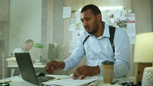 Serious detective opening folder with criminal records, homicide case files and typing on laptop sitting at police office. Policeman doing paper work, filling reports and documents on computer. 4K