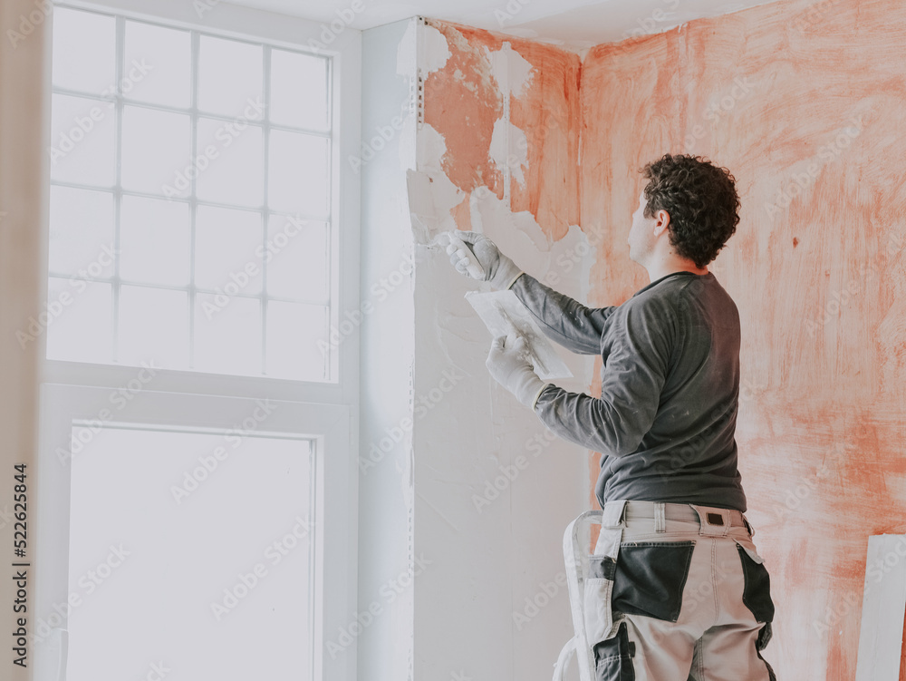 A young caucasian guy applies fresh putty with a small spatula on the wall near the window A young caucasian guy applies fresh putty with a small spatula on the wall near the window