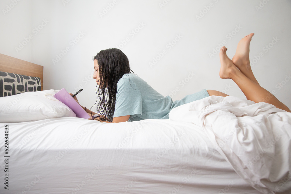Caucasian woman wearing only a t-shirt writing in her notebook on the bed Caucasian woman wearing only a t-shirt writing in her notebook on the bed