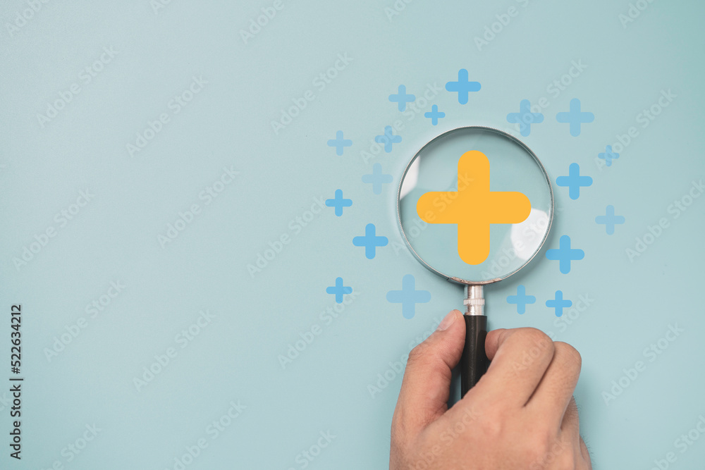 Hand holding magnifier glass with yellow plus sign symbol inside for focus healthcare insurance and offer positive thinking mindset of personal development concept. - obrazy, fototapety, plakaty 