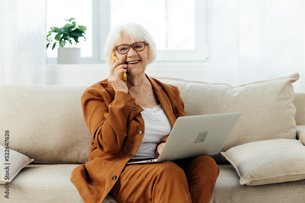 a happy elderly woman smiles sweetly sitting on the sofa in a bright apartment near the window and talking on a smartphone holding a laptop on her lap a happy elderly woman smiles sweetly sitting on the sofa in a bright apartment near the window and talking on a smartphone holding a laptop on her lap