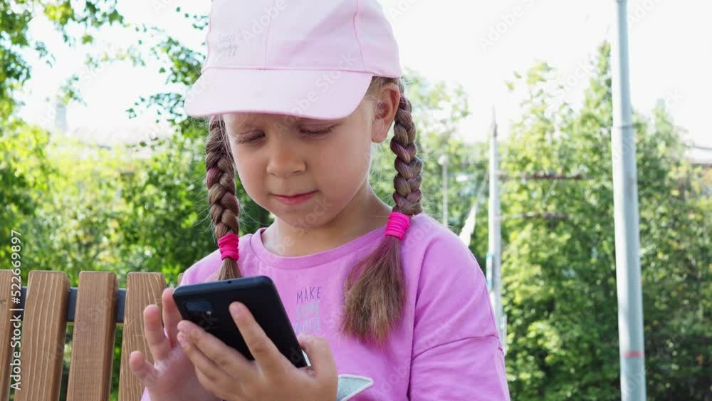 A small, beautiful preschool girl sits on a bench in the park in summer and plays on the phone. A child in protection on his knees with a skateboard with a phone in his hands. A small, beautiful preschool girl sits on a bench in the park in summer and plays on the phone. A child in protection on his knees with a skateboard with a phone in his hands.