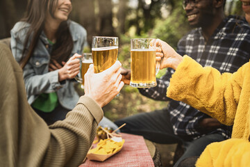 A cheerful group of multiracial friends relax while toasting while sitting in the pub in the woods and drinking ice-cold beers - detail on hands with mugs raised