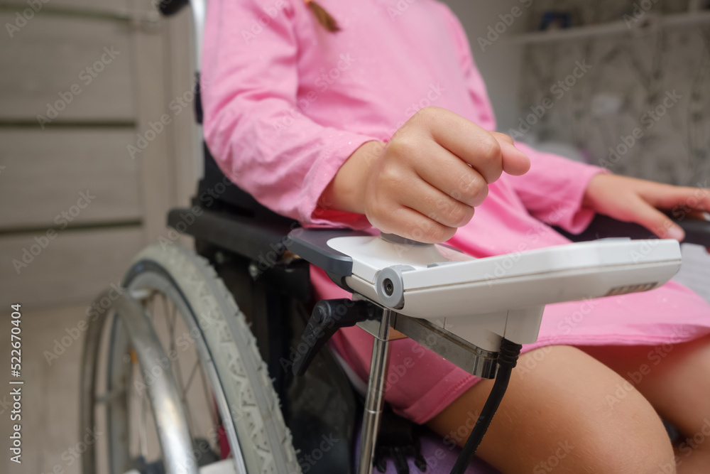 Obraz premium baby girl in a dress sitting on an electric wheelchair indoors. close-up photo of electric wheelchair joystick
