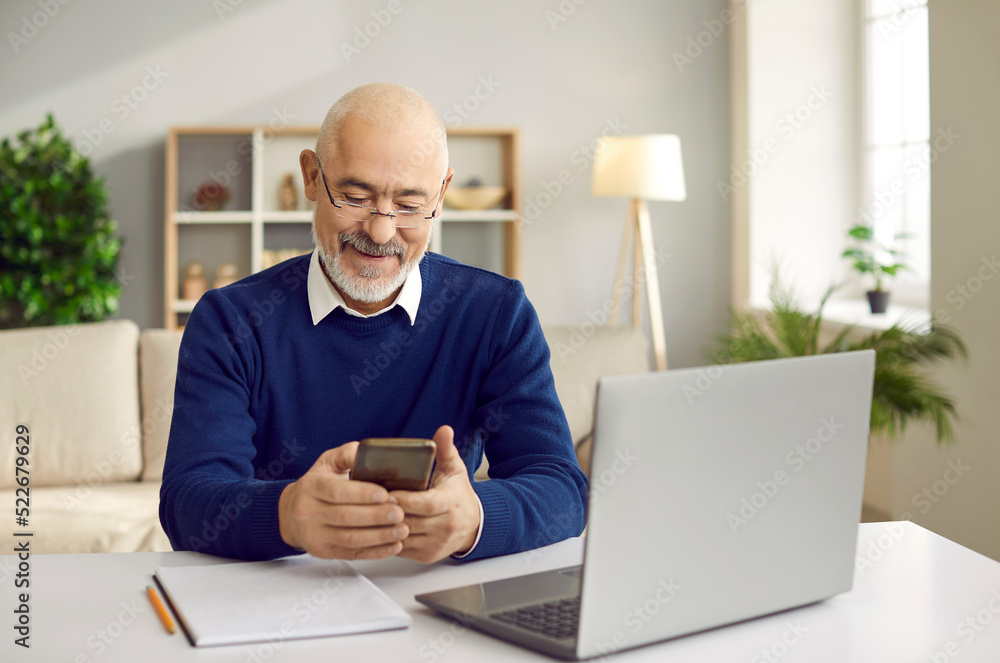 Senior man using mobile phone. Happy retired man in glasses sitting at desk with laptop and cellphone, paying bills online, reading news, sending emails, making banking transaction, calling his family Senior man using mobile phone. Happy retired man in glasses sitting at desk with laptop and cellphone, paying bills online, reading news, sending emails, making banking transaction, calling his family
