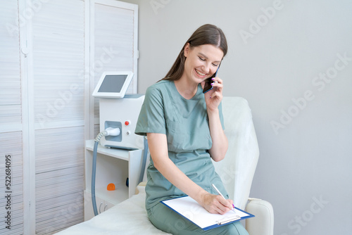 Cheerful woman doctor in green coat in laser hair removal room records clients for a procedure on the phone