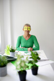 Middle-aged woman with short hair and green top is sitting at office table with many green plants and working notes and has sticky notes with painted eyes on her face