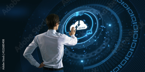 Back view of young businessman pointing at abstract glowing cloud hologram on blurry dark background. Cloud computing, big data backup concept. photo