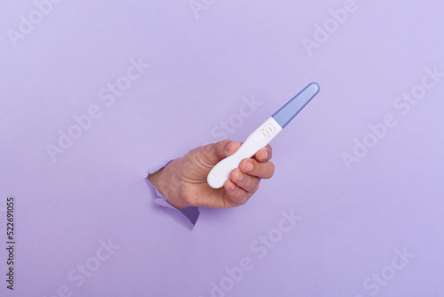 person's hand holding pregnant test in torn hole of purple background, medical healthcare gynecological, pregnancy, fertility, maternity. Copy space for advertisement text inscription.