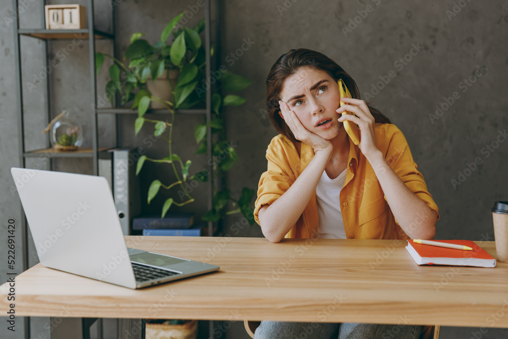 Young sad tired minded employee business woman 20s wearing casual yellow shirt talk speak on mobile cell phone put look aside sit work at wooden office desk with pc laptop Achievement career concept. Young sad tired minded employee business woman 20s wearing casual yellow shirt talk speak on mobile cell phone put look aside sit work at wooden office desk with pc laptop Achievement career concept.